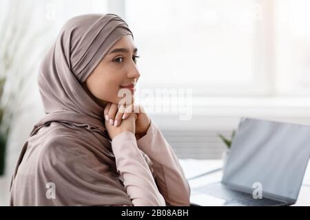 Lächelnde muslimische Geschäftsfrau, die Kopftuch trägt, am Arbeitsplatz sitzt und wegschaut Stockfoto