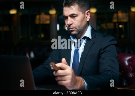 Geschäftsmann, der in einer Bar im Business Center sitzt und Whisky trinkt, der Zigarre bekommt Stockfoto