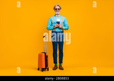 Großes Foto von coolen Opa Hold Tickets empfehlen Reisen mit dem Flugzeug Rollender Koffer tragen Sie blaue Hemd Hosenträger Bow Krawatte Hosen Stiefel Socken Stockfoto