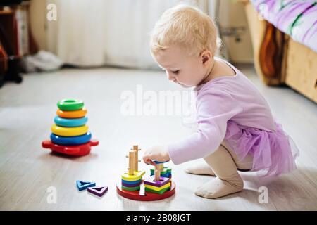 Mädchen spielt logische Pyramide auf dem Boden im Wohnzimmer am sonnigen Tag. Montessori Holzspielzeug gefaltete Pyramide. Mehrfarbiges Spielzeug blau, gelb, rot, grün. Stockfoto