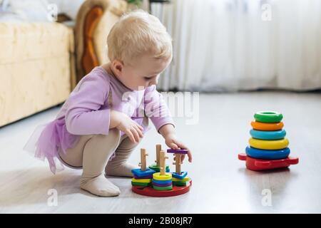 Mädchen spielt logische Pyramide auf dem Boden im Wohnzimmer am sonnigen Tag. Montessori Holzspielzeug gefaltete Pyramide. Kreis, Quadra, Dreieck, rechteckiges Holzelem Stockfoto