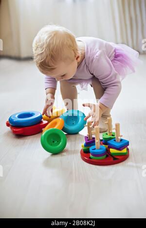 Das Mädchen hebt die Pyramidenringe aus der weißen Hülse im Wohnzimmer. Montessori Holzspielzeug gefaltete Pyramide. Kreis, Quadra, Dreieck, rechteckige Holzelemente von c Stockfoto