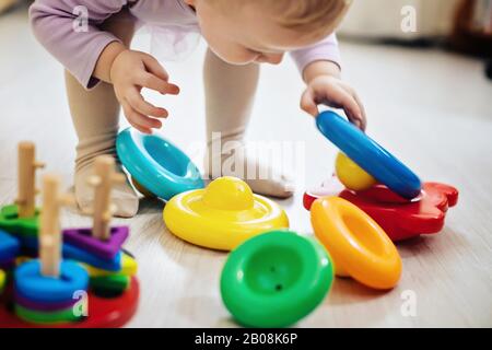 Das Mädchen hebt die Pyramidenringe aus der weißen Hülse im Wohnzimmer. Montessori Holzspielzeug gefaltete Pyramide. Mehrfarbiges Spielzeug blau, gelb, rot, grün. Stockfoto