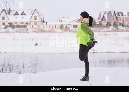 Seitenansicht der jungen Frau mit schwarzem Haar, das sich mit dem Bein ausdehnt, während Sie sich auf das laufen im Winterpark vorbereiten Stockfoto