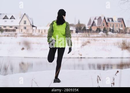 Rückansicht der brünettigen Frau, die eine vierfache Dehnübung macht, während sie sich das Bein an der Winterflußküste streckt Stockfoto