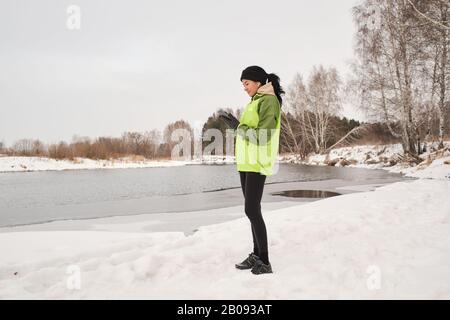 Junge brünette Frau mit Pferdeschwanz, die am Fluss steht und die Laufstrecke auf dem Smartphone analysiert Stockfoto