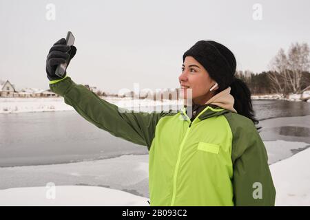Positive schöne Frau in Ohrenknospen, die an der Winterküste steht und sich auf der Smartphone-Kamera fotografiert Stockfoto