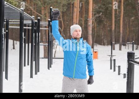 Gutaussehender junger Mann in blauer Jacke, der im Winter auf dem Sportplatz trainieren kann, während er Bewegung auf der Schulter macht Stockfoto