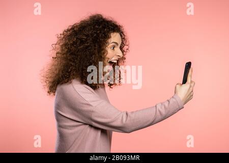 Wütende geschweifte Frau schreit herab, schwört mit jemandem. Verwenden des Mobiltelefons. Gestresste und depressive Mädchen auf pinkfarbenem Hintergrund. Stockfoto