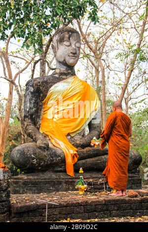 Ein buddhistischer Mönch in orangefarbenem Kleid betet vor einer alten Statue Stockfoto