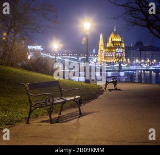 Budapest, Ungarn - Bank- und Lampenposten in einem Park im Buda-Viertel mit Szechenyi-Kettenbrücke und Parlament im Hintergrund zur Winterzeit Stockfoto