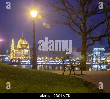 Budapest, Ungarn - Bank- und Lampenposten in einem Park im Buda-Viertel mit Szechenyi-Kettenbrücke und Parlament im Hintergrund zur Winterzeit Stockfoto