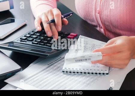 Die Buchhalterin im Büro hält einen Kassierercheck in den Händen und erstellt Berechnungen auf einem Rechner, wobei sie die Ergebnisse in einem Notizbuch aufschreibt. Stockfoto