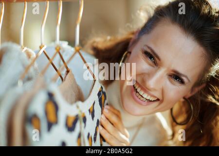 Porträt der glücklichen jungen Frau in weißem Pullover und Rock im modernen Haus am sonnigen Wintertag, die Pullover an einem kupfernen Kleiderständer hängen. Stockfoto