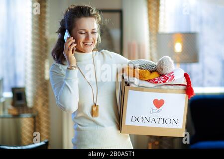Fröhliche, stilvolle Frau in weißem Pullover und Rock mit Spenderbox mit alter warmer Kleidung, die im sonnigen Wintertag auf einem Handy zu Hause spricht. Stockfoto