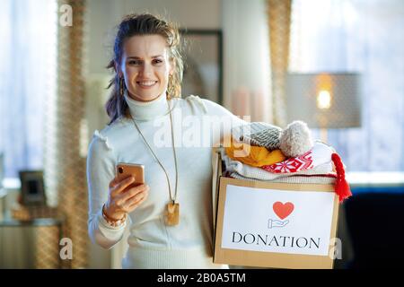 Fröhliche, stilvolle Hausfrau im mittleren Alter in weißem Pullover und Rock mit Spenderbox mit alter warmer Kleidung mit App auf dem Smartphone im modernen Wohn-Roo Stockfoto