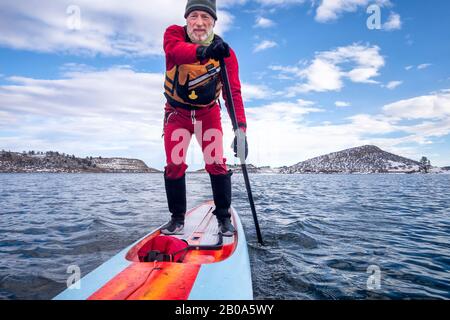 Senior-Paddler in einem Trockenanzug und einer Schwimmweste paddeln im Winter auf einem See in Colorado - Horset ein langes, unbegrenztes Stehpaddleboard Stockfoto