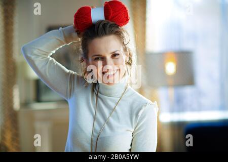 Porträt einer lächelnden jungen Frau in weißem Pullover und Rock, die sich im modernen Wohnzimmer im sonnigen Wintertag mit roten Strickgarnhosen verbeugen. Stockfoto