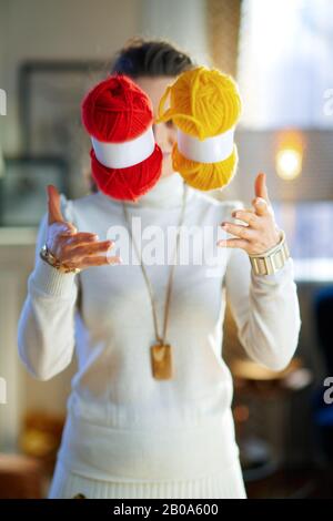 Nahaufnahme der jungen Frau im weißen Pullover und Rock im modernen Haus am sonnigen Wintertag, die gelbe und rote Strickgarnhosen aufwirft. Stockfoto