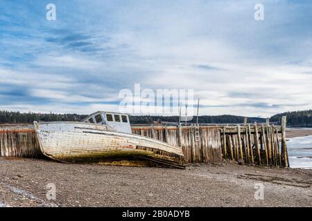 Ein verunglücktes Boot am Ufer neben einem verfallenen Kai. Sowohl Boot als auch Wharf sind in sehr schlechter Form und fallen auseinander. Ebbe. Stockfoto