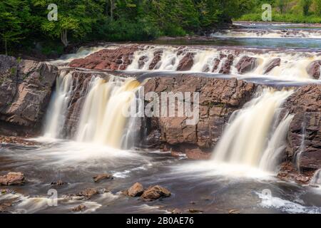 Lepreau Falls in New Brunswick, Kanada. Lange Belichtung erweicht den Wasserfall, wenn er über braunem Felsen fließt. Bäume am Fluss, sonniger Tag. Stockfoto