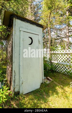 Ein weißes Outhouse neben einem kleinen weißen Holzzaun. In der Tür ist ein Halbmond hoch geschnitten. Gras davor und Bäume dahinter. Stockfoto