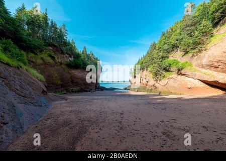 Ein Strand zwischen zwei Klippen am Meer. Eine Klippe ist in hellem Sonnenlicht, die andere ist im Schatten, ebenso wie der Großteil des Strandes. Blauer Himmel und blauer Ozean. Stockfoto