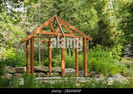 Braun gefärbtes Holzholz und Holzpergola auf Betonplatten, die im Sommer inmitten von bewaldeten Gärten von großen Natursteinen umgeben sind. Stockfoto