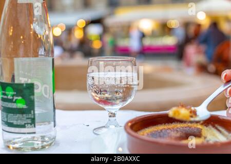 Selektiver Fokusschuss aus einem Glas und einer Flasche kohlensäurehaltiges Wasser zusammen mit einem Löffel cremefarbenen Brulee in einem Outdoor-Café in der Nacht. Stockfoto