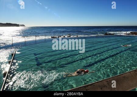 Sydney, Australien, am Bronte Ocean Pool, ein Schwimmschwimmer in einer rosafarbenen Kappe, das grüne Wasser wackelt am Morgen vom Sonnenlicht, während Surfer auf die Wellen warten Stockfoto