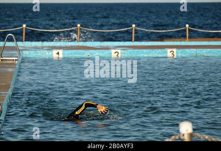 Ein einsamer Mann schwimmt in einem Wetsuit mit Handpaddles in einem Ozeanbecken bei Dee Why an Sydneys North Shore, enger horizontaler Bildröber auf der linken Seite Stockfoto