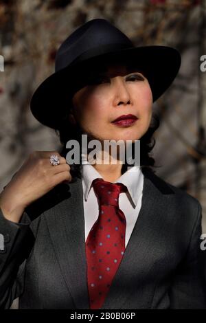 Gesicht im teilweisen Schatten, eine Asianerin, die eine Sharkskin-Suite im Stil eines Mannes trägt, rote Krawatte und Fedora-Hut, die wie ein Gangster der vierziger Jahre aussehen, rote Lippen Stockfoto