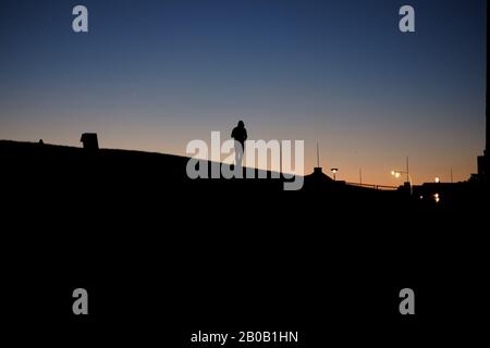 Silhouette von Männern, die in der Dämmerung auf der Kurve eines Hügels spazieren gehen, Straßenbeleuchtung, klarer Himmel, blaues und orangefarbenes Leuchten am Horizont, das eine Kapuze trägt Stockfoto