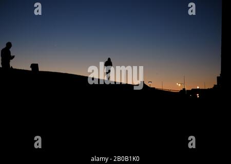 Silhouette von Männern, die in der Dämmerung auf der Kurve eines Hügels spazieren gehen, Straßenbeleuchtung, klarer Himmel, blaues und orangefarbenes Leuchten am Horizont, das eine Kapuze trägt Stockfoto