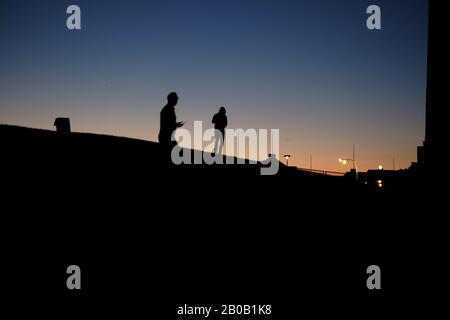 Silhouette von Männern, die in der Dämmerung auf der Kurve eines Hügels spazieren gehen, Straßenbeleuchtung, klarer Himmel, blaues und orangefarbenes Leuchten am Horizont, das eine Kapuze trägt Stockfoto