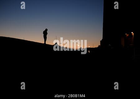 Silhouette von Männern, die in der Dämmerung oder im Morgengrauen auf der Kurve eines Hügels spazieren gehen, Stretlights, klarer Himmel, blaues und orangefarbenes Leuchten am Horizont, das eine Kapuze trägt Stockfoto