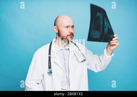 Attraktiver freundlicher kahler lächelnder Arzt, der auf blauem Hintergrund isolierte Röntgenaufnahmen ansieht Stockfoto