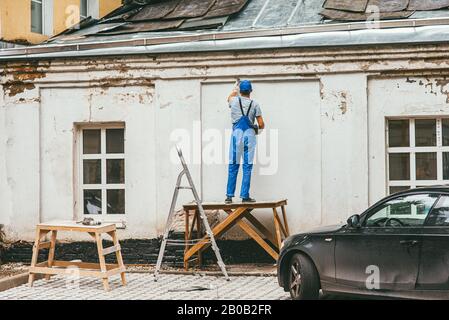 Nicht erkennbarer Arbeiter in blauer Bauhose steht auf einem Holzständer und arbeitet mit einem Spachtel mit Gips an der Wand. Renovierung der Stockfoto