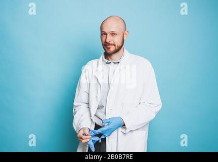 Attraktiver lächelnder Mann kahler bärtiger Arzt legt Gummihandschuhe an, die auf dem blauen Hintergrund isoliert auf die Kamera blicken Stockfoto