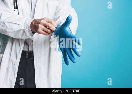 Erntefoto des Arztes in weißem Mantel legt auf Gummihandschuhe, die auf dem blauen Hintergrund isoliert sind Stockfoto