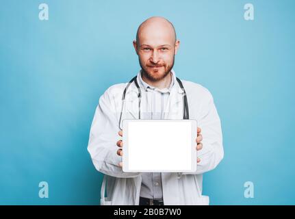 Der attraktive, freundlich kahle lächelnde Arzt hält Tablette mit weißem Bildschirm, der auf die Kamera blickt, die auf dem blauen Hintergrund isoliert ist Stockfoto