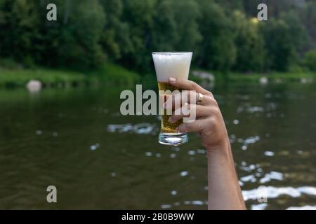 Glaskoblet mit Bier in hoher Hand vor dem Hintergrund der Sommerlandschaft. Nahaufnahme des Mannes, der ein Glas Bier in der Hand hält. Stockfoto