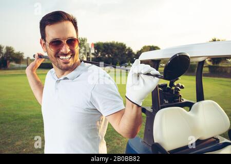 Ein gutaussehender junger Mann hält seinen Golfclub vor der Golfkarre. Stockfoto