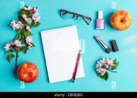 Federtürkisfarbenes Layout mit weißem Notizblock mit Stift, Frauenbrille, pinkfarbenem Lippenstift und nagellack, zwei Äpfeln und pinkfarbenen Apfelbaumzweigen. Frau zu Hause Stockfoto