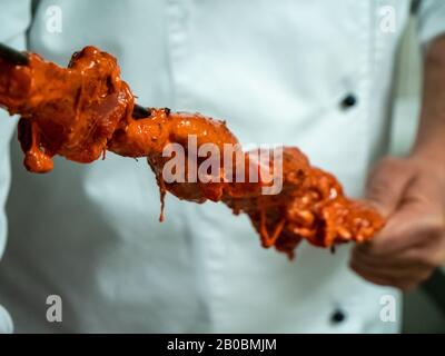 Ein indischer Koch schiebt mariniertes Huhn auf einen langen Metallküchespieß, der für eine Tandoori-Küche bereit ist. Stockfoto