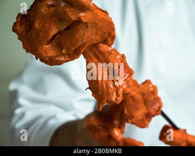 Ein indischer Koch schiebt mariniertes Huhn auf einen langen Metallküchespieß, der für eine Tandoori-Küche bereit ist. Stockfoto