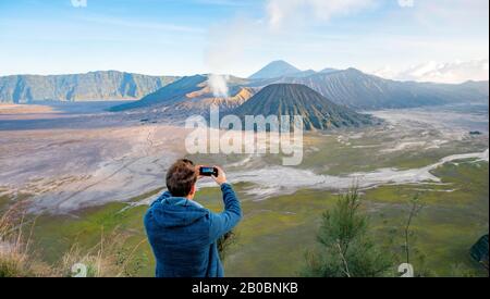 Junger Mann fotografieren mit iPhone, Vulkanlandschaft, Blick in Tengger Caldera, rauchender Vulkan Gunung Bromo, vor Mt. Batok, im Rücken Mt. Stockfoto