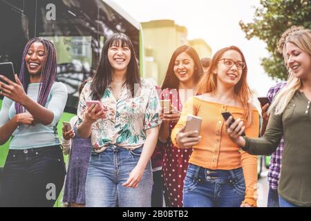 Fröhliche Freunde, die Smartphones am Busbahnhof nutzen - Junge Studenten, die nach der Schule im Freien mit Technologietrends Spaß haben - Freundschaft, Universität Stockfoto