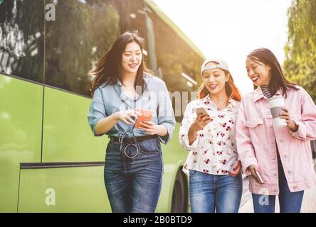 Fröhliche asiatische Freunde, die Smartphones am Busbahnhof nutzen - Junge Studenten, die sich nach der Schule im Freien über Technologietrends lustig machen - Freundschaft, Univ Stockfoto