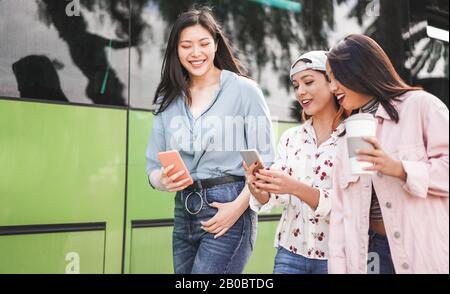 Fröhliche asiatische Freunde, die Smartphones am Busbahnhof nutzen - Junge Studenten haben nach der Schule Spaß mit Technologietrends - Freundschaft und Transport Stockfoto